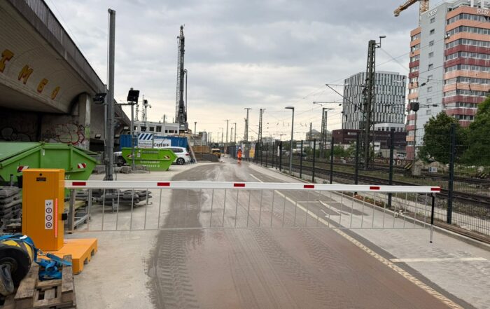 Mobile Schranke vor einer Baustelle, im Hintergrund Bahngleise und Industrie
