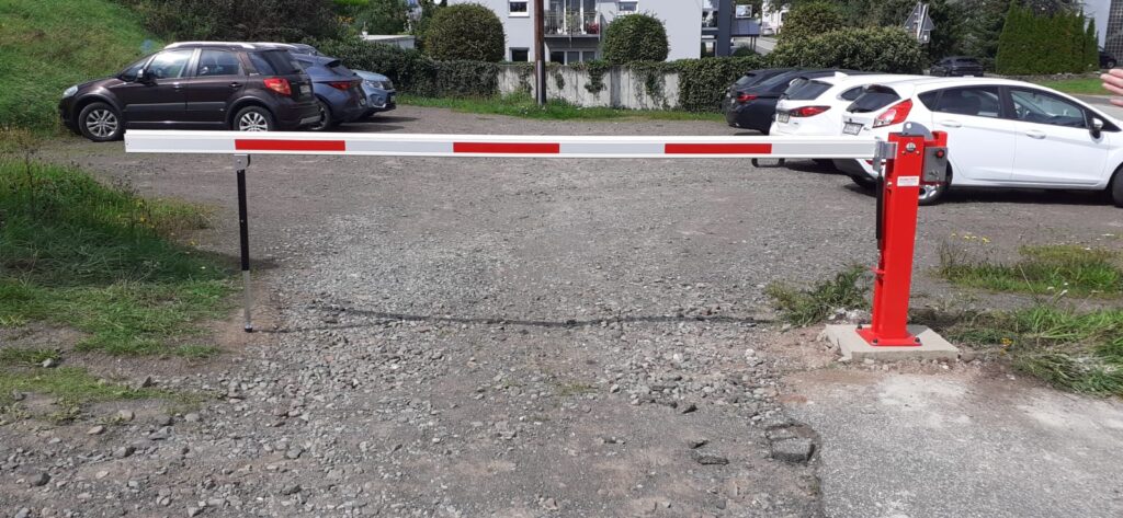 Rot-weiße Schranke an einer Schotterzufahrt mit parkenden Fahrzeugen im Hintergrund.