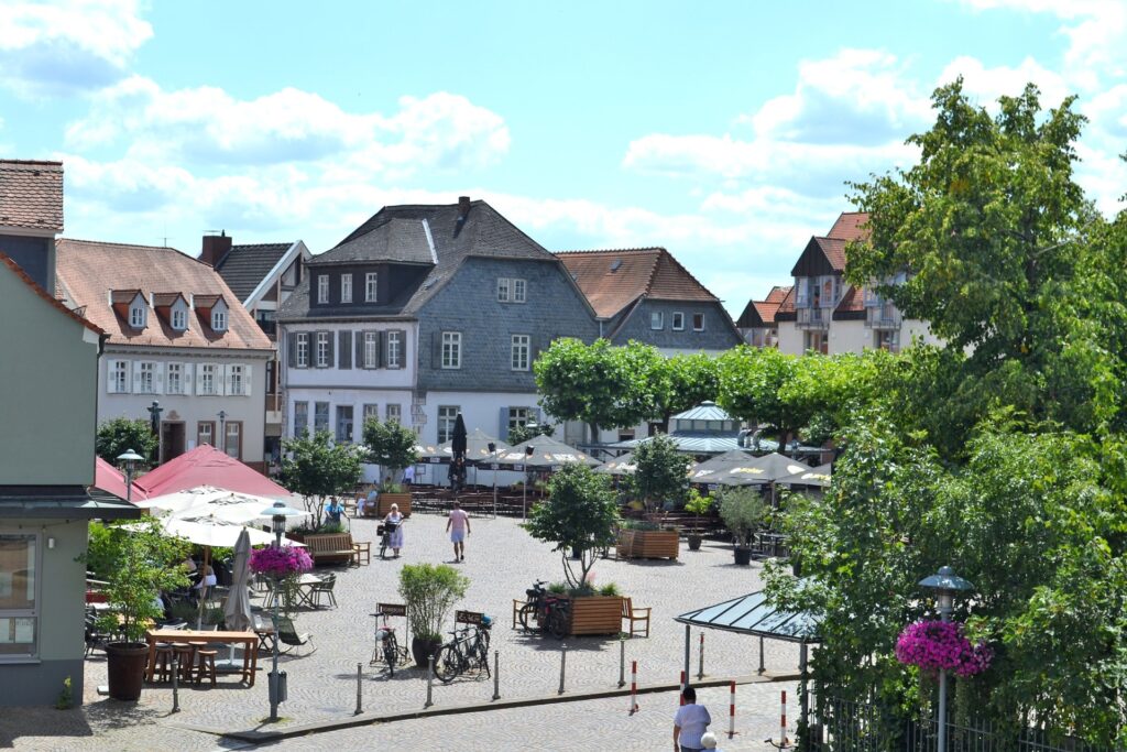 Belebter Innenstadtplatz mit Gastronomie im Freien, Bäumen und niedrigen Absperrpollern am Rand des Kopfsteinpflasters.