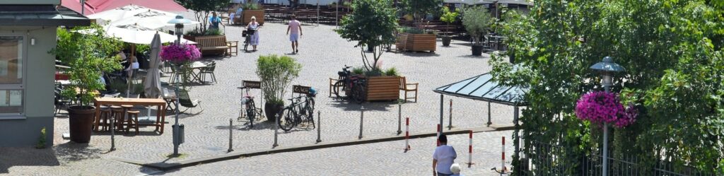 Belebter Innenstadtplatz mit Gastronomie im Freien, Bäumen und niedrigen Absperrpollern am Rand des Kopfsteinpflasters.