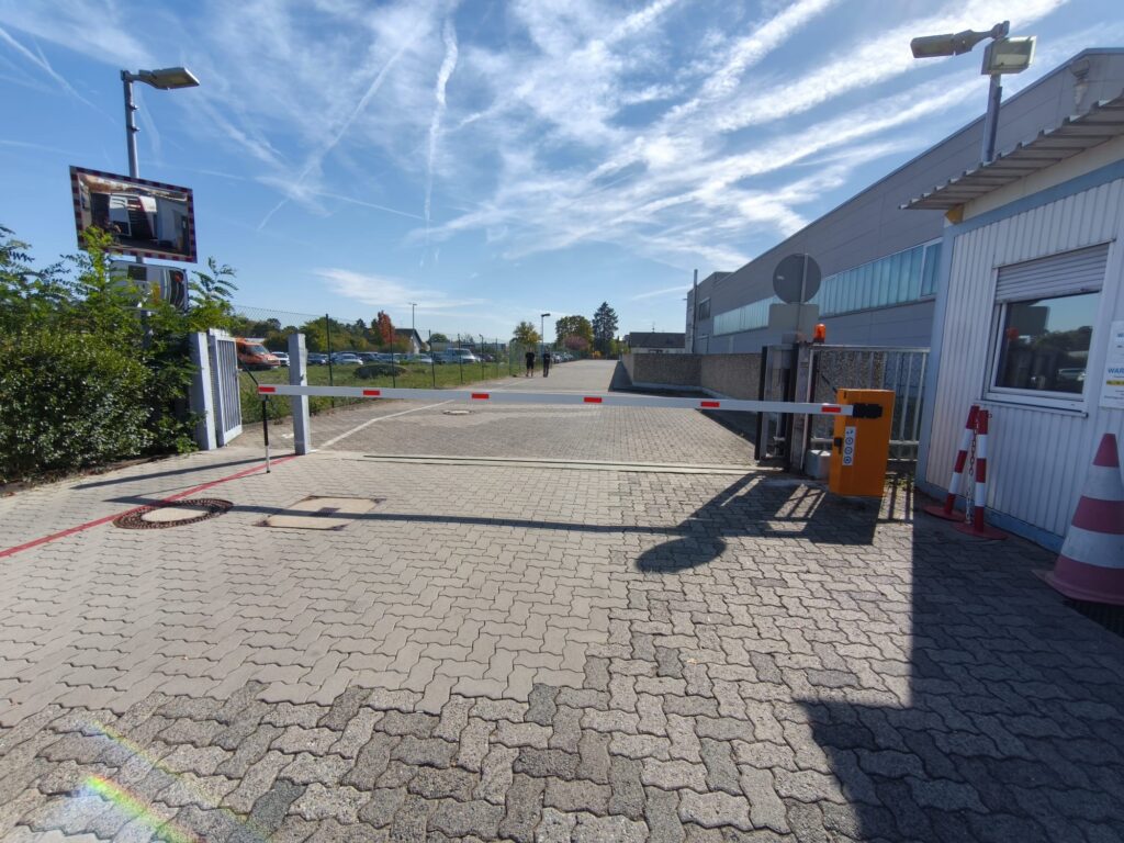 Gelb-orange Schranke auf gepflasterter Zufahrt, dahinter ein Betriebsgelände, rechts ein Pförtnerhäuschen