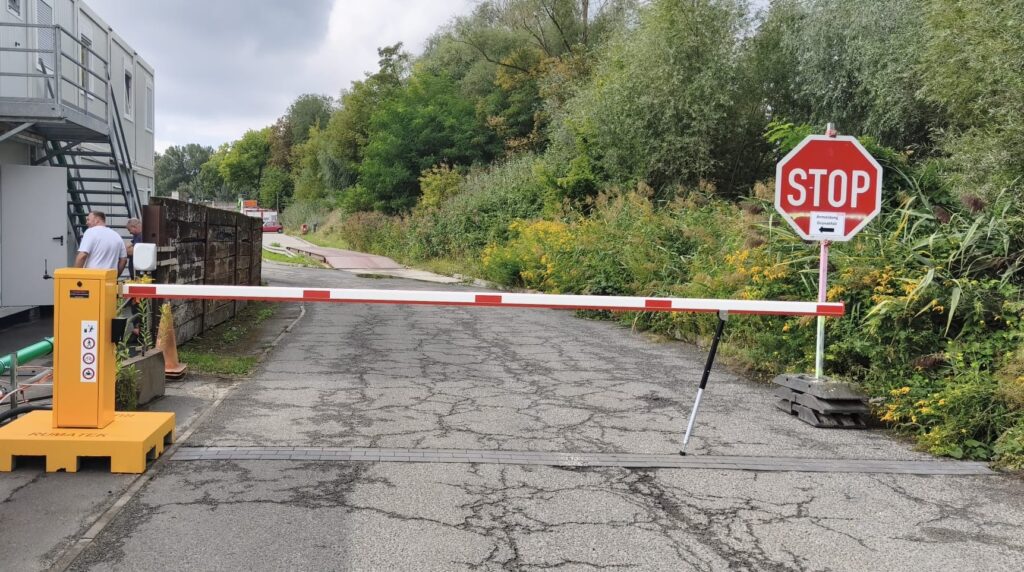Mobile Schranke in orange vor einer Zufahrt, daneben Container