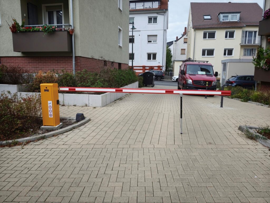 Eine Schranke mit weiß-rotem Schrankenbaum vor einer geplasterten Zufahrt, dahinter ein Kleintransporter